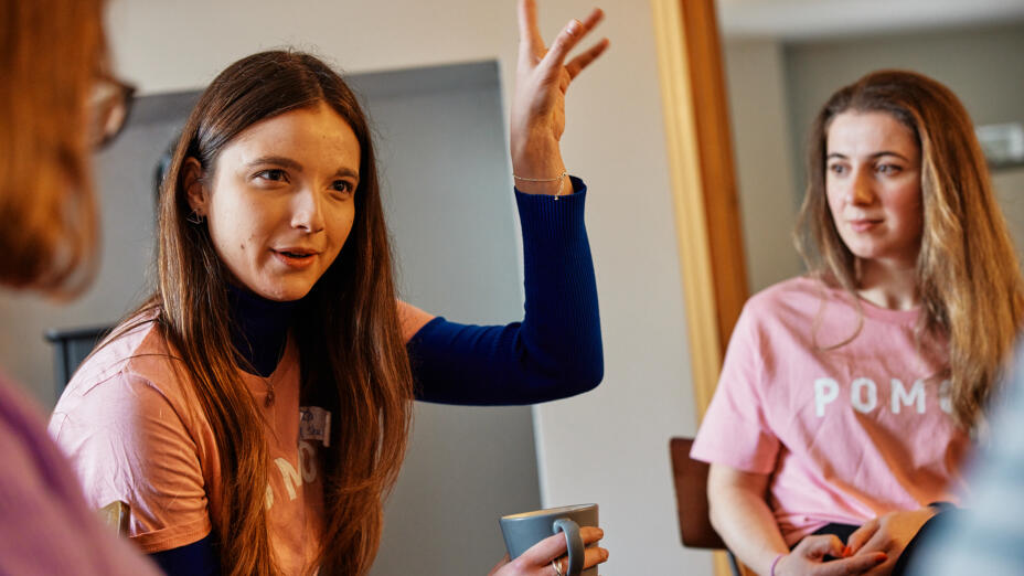 Two young women in pink POMOC t-shirts in conversation at a residential, one with her hand raised mid-sentence.